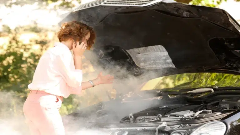 A stressed woman talking on the phone next to a smoking car engine with an open hood, highlighting the need for a trusted european car workshop